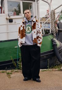 Ovrhnjen plašč iz rjavega in belega umetnega krzna, siva kapucar s krožnim logotipom, črne široke hlače in črni čevlji. V ozadju je zelen čoln.