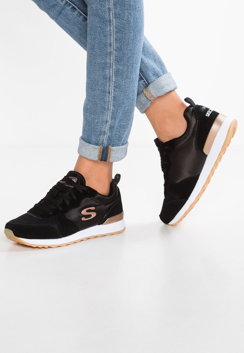 Schwarze Sportschuhe mit Obermaterial aus Mesh und Wildleder, roségoldenen Akzenten, weißer Zwischensohle und Gummisohle; kombiniert mit hochgekrempelten blauen Jeans.