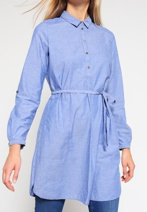 Jasnoniebieska sukienka koszulowa z długim rękawem, zapięciem na guziki przy kołnierzyku, paskiem w talii i przednimi kieszeniami, noszona z niebieskimi dżinsami.