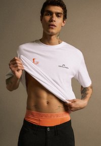 Biała bawełniana koszulka z czerwoną grafiką surferską i czarnym logo. Uzupełniona pomarańczowym pasem z napisem "COALITION". Jednolite ciemne spodnie.