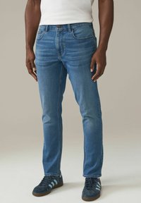 Hombre con jeans ajustados de color azul medio, camiseta blanca metida por dentro y zapatillas oscuras con rayas azul claro, de pie frente a un fondo liso.