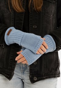 Gants sans doigts en tricot bleu clair, avec une texture côtelée et une étiquette en cuir marron. Portés sur une veste noire et un jean.