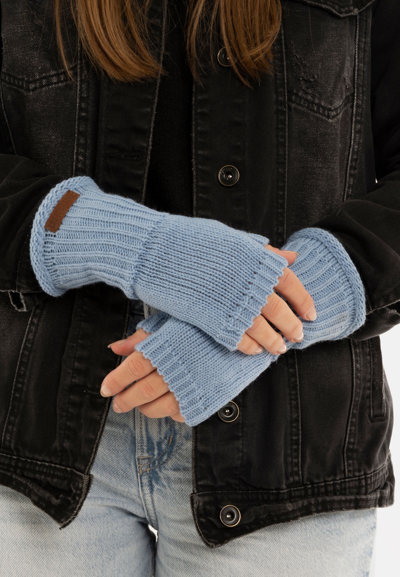 Gants sans doigts en tricot bleu clair, avec une texture côtelée et une étiquette en cuir marron. Portés sur une veste noire et un jean.