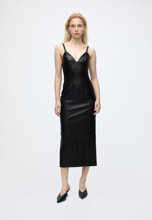 Femme aux cheveux blonds portant une robe midi noire sans manches avec de fines bretelles et des talons noirs pointus, debout sur un fond blanc uni.