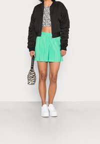 Sort bomberjakke over en zebra-mønstret crop top, parret med plisserede grønne shorts, hvide sneakers og en lille stribet håndtaske.
