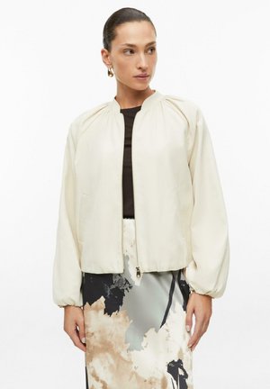 Femme aux cheveux foncés tirés en arrière porte une veste beige clair zippée sur un top noir et une jupe taille haute à imprimé abstrait beige et noir.