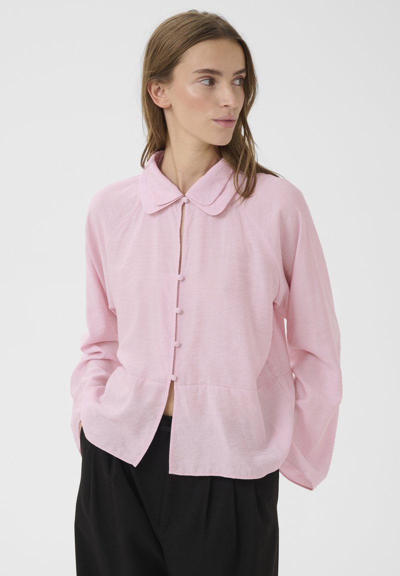 Naine kannab lahtist heleroosat nööpidega pluusi ümarate kraega ja musti pükse, vaatab küljele ning hoiab käed taskutes.