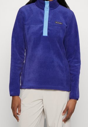 Kvinde iført en langærmet lilla fleece trøje med lyseblåt knapstykke og beige bukser, stående mod en hvid baggrund.