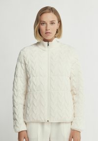 Vit stickad jacka med hög krage, texturerat kabelmönster, hel dragkedja och något puffy silhuett, designad för värme och komfort.