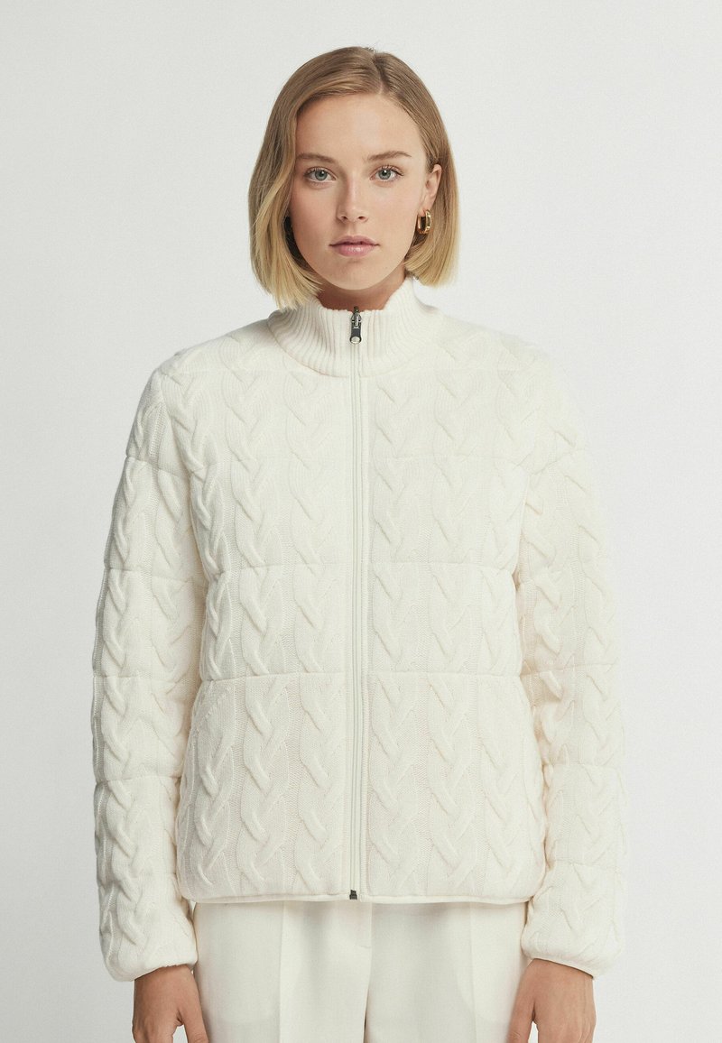 Vit stickad jacka med hög krage, texturerat kabelmönster, hel dragkedja och något puffy silhuett, designad för värme och komfort.