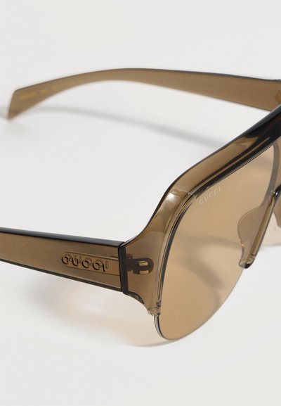 Καφέ διαφανή υπερμεγέθη γυαλιά ηλίου με φαρδιές βραχίονες που φέρουν το λογότυπο Gucci στο πλάι, στρογγυλεμένους φακούς και ενσωματωμένα ρινικά μαξιλαράκια.
