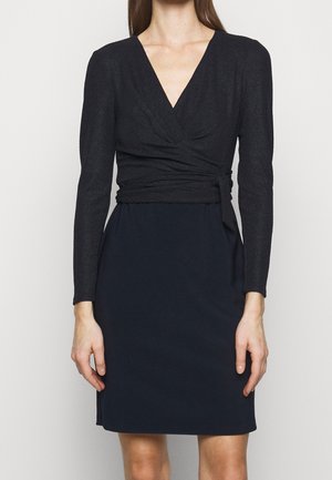 Vrouw draagt een zwarte lange-mouwen wikkeltop en een marineblauwe knie-lange rok tegen een effen lichte achtergrond.