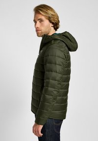 Olivgrön puffjacka med en passande huva, sid sömmar och quiltat mönster. Tillverkad av ett slätt, lätt vikt material.