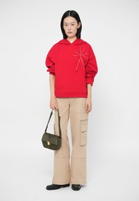 Κόκκινο φούτερ με κουκούλα και σχεδιασμό φιόγκου, συνδυασμένο με μπεζ παντελόνι cargo. Το μοντέλο κρατάει μια πράσινη τσάντα χειρός. Λείο ύφασμα, άνετη εφαρμογή, φαρδύ παντελόνι.