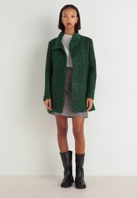 Manteau en mélange de laine vert à finition texturée, design croisé, avec des boutons noirs, porté sur un haut gris et une jupe, associé à des bottes noires.