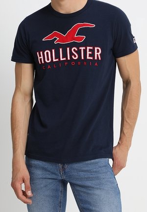 Homme portant un t-shirt bleu marine Hollister California avec un logo rouge et un jean bleu clair, debout avec une main dans la poche.