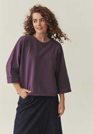 Ženska z kodrastimi lasmi, oblečena v vijoličasto ohlapno majico in temno krilo, pogleda vstran z rahlim nasmeškom, roka v žepu.