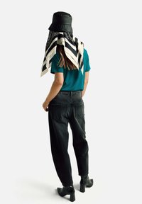 Osoba v tyrkysovém tričku, černých džínách s vysokým pasem, černých botách na podpatku, černém klobouku ve tvaru kbelíku, se šálou s černobílým vzorem visící na zádech.