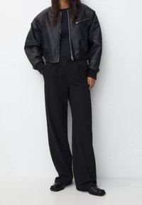 Schwarze Lederbomberjacke mit gerippten Bündchen, kombiniert mit hoch taillierten schwarzen Hosen. Die Schuhe zeichnen sich durch ein glattes, minimalistisches Design aus.