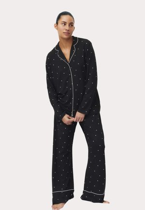 Vrouw staat op blote voeten en draagt een zwarte pyjama met lange mouwen, versierd met kleine witte hartjes en witte biesdetails.