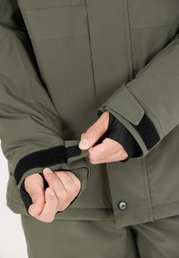 Grøn vandtæt jakke med justerbare cuffs. Ærmerne har sorte Velcro-stropper og et tætsiddende indvendigt foder for ekstra beskyttelse.