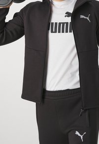 Chaqueta deportiva negra con cremallera y bolsillos laterales, combinada con una camiseta gris con el logo de PUMA. Pantalones de chándal negros con detalle del logo.