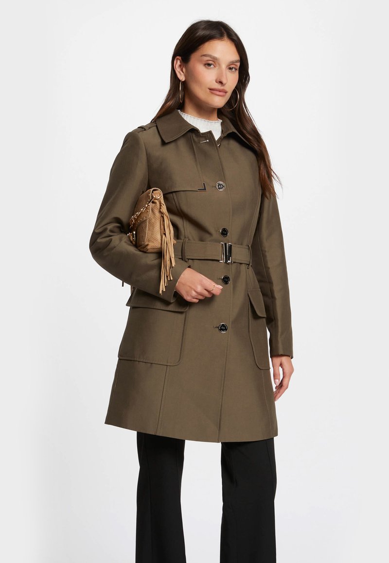 Olijf groene trenchcoat met een ceintuur in de taille, knoopsluiting en klepzakken. Combinatie met een gestructureerde beige handtas en een crèmekleurige trui.