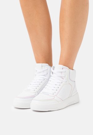 Baskets montantes blanches à coutures multicolores portées sur des jambes nues, présentées sur un fond clair uni.