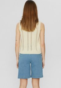 Gebreide mouwloze top in crème, met verticale opengebreide patronen, gecombineerd met lichtblauwe denim shorts met een rechte snit en omgeslagen pijpen.