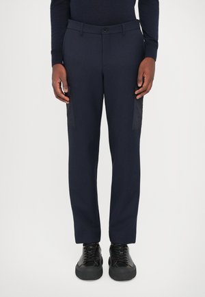 Marineblaue Hose mit taillierter Passform, aus glattem Stoff, mit seitlichen Cargo-Taschen in Schwarz und Knopfverschluss vorne, kombiniert mit schwarzen Sneakers.