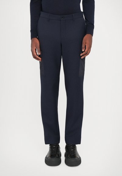 Pantalones azul marino con un corte ajustado, que presentan una tela suave, bolsillos laterales tipo cargo en negro y cierre frontal con botón, combinados con zapatillas negras.