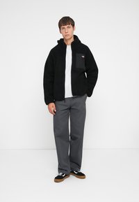 Veste en polaire noire avec une poche zippée sur la poitrine, portée sur un t-shirt gris, associée à un pantalon gris et des chaussures noires avec des accents blancs.