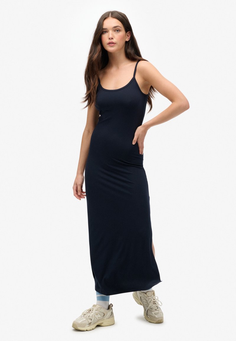 Robe maxi bleu marine avec des bretelles spaghetti, forme ajustée, tissu doux, fentes sur les côtés, associée à des baskets de sport de couleur claire.