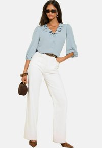 Femme portant un chemisier à volants bleu clair, un pantalon large taille haute blanc, des talons pointus marron et des lunettes de soleil, tenant un sac à main tissé marron.
