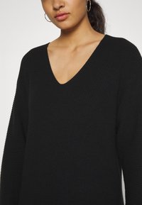 Pull en tricot noir avec un décolleté en V profond, texture côtelée et manches longues. Le tissu semble doux et légèrement épais. Les détails incluent un encadrement au niveau de l'encolure.