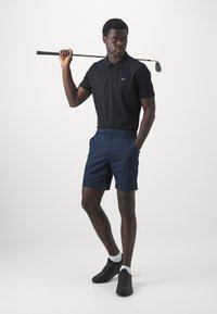 Črna majica s polo ovratnikom s CK logotipom, mornarske kratke hlače in črne čevlje. Golf palica resting na rami. Bela podlaga poudarja oblačila.