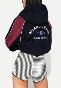 Woman facing away wearing a black and burgundy hooded jacket with "Atlantic Miles" text and striped black-and-white shorts.