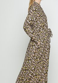 Nainen sivuprofiilissa, päällä pitkä, kukkakuosiset mekko, jossa on vaaleanpunaisia, keltaisia ja valkoisia kukkia, kädet taskuissa yksinkertaisen taustan edessä.