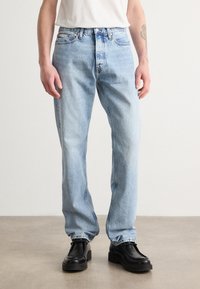 Vaqueros de mezclilla de color azul claro con un ajuste holgado y pierna recta. Presenta un diseño estándar de cinco bolsillos y costuras en contraste. Llevados con zapatos negros.