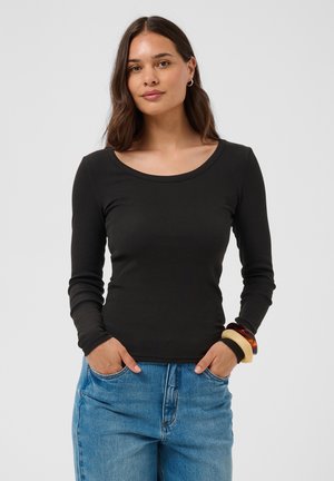 Jeune femme aux longs cheveux foncés porte un haut noir ajusté à manches longues, un jean bleu et des bracelets épais, debout avec les mains dans les poches.