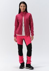 Fleece-Zip-Jacke in leuchtendem Pink, kombiniert mit pinken und schwarzen Stretchpants. Beide zeichnen sich durch ein elegantes Design und kontrastierende Akzente aus. Dunkles Schuhwerk rundet den Look ab.