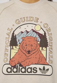 Krämsvit sweatshirt med en björnteckning, berg och träd. Tryckt text läser "OFFICIELL GUIDE ORIGINALS" och "adidas" logotyp.