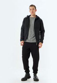 Sort softshell-jakke med hætte, grå T-shirt og sorte sweatpants. Tyk tekstur, lynlåslommer og kraftige sorte støvler.