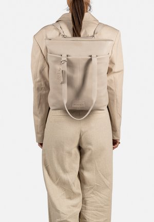 Beige leren rugzak met een gestructureerd ontwerp, bovenflap en verstelbare banden. Heeft een label en logo aan de voorkant.