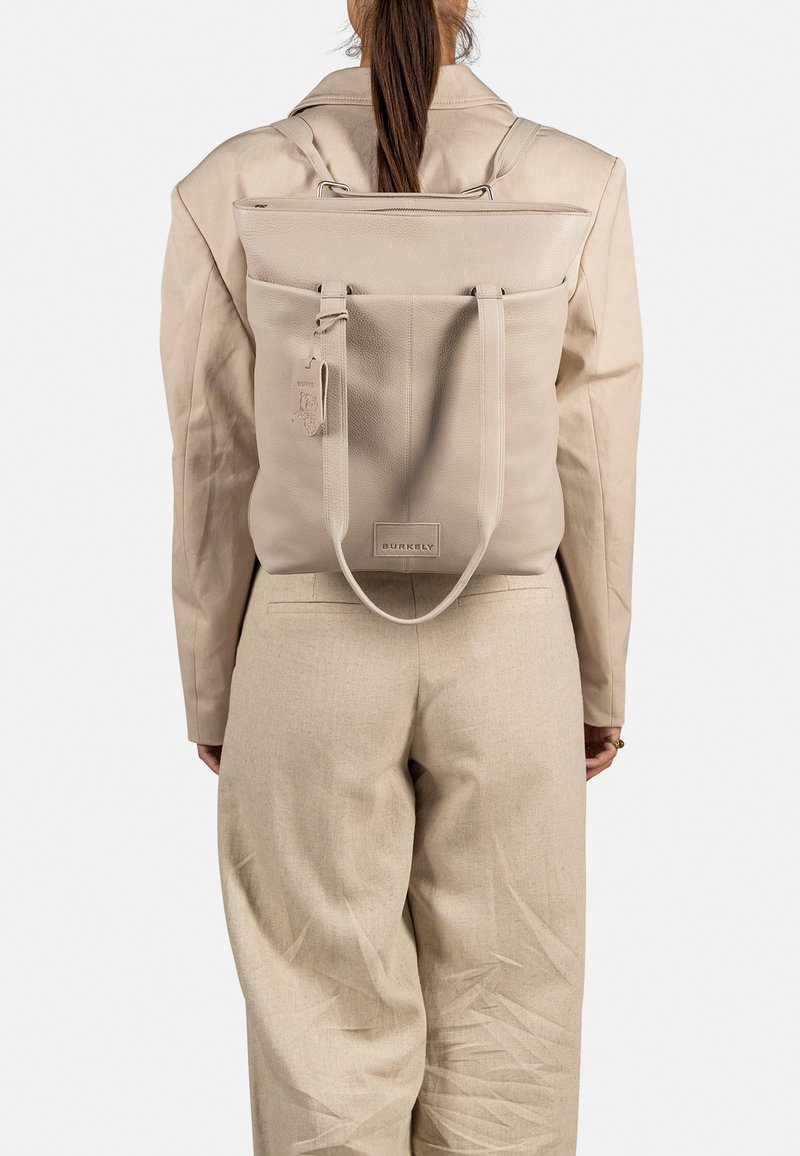 Zaino in pelle beige con un design strutturato, pattina superiore e spallacci regolabili. Presenta un'etichetta e un logo sul davanti.