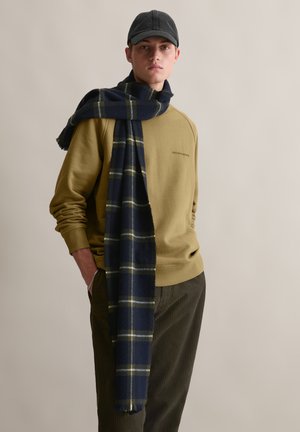 Olive green sweatshirt with a crew neckline, paired with dark green corduroy pants. Dark plaid scarf with blue and yellow accents draped casually.