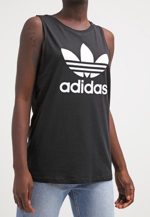 Osoba nosząca czarną bezrękawnikową koszulkę Adidas z białym logo i jasnoniebieskie dżinsy, stojąca na jednolitym białym tle.