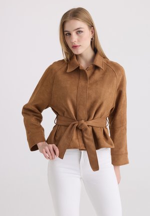 Young woman with straight blonde hair wearing a brown suede belted jacket and white pants, facing forward against plain background.