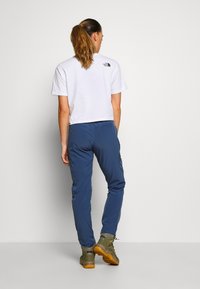 T-shirt blanche coupée avec un petit logo au dos, associée à un pantalon bleu et des bottes de randonnée marron. Tissu lisse, design décontracté.