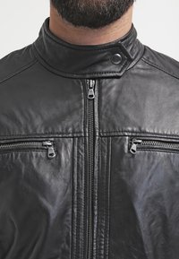 Černá kožená bunda má vysoký límec, přední zip a dvě zipové kapsy na hrudi. Textura je hladká s mírným leskem.